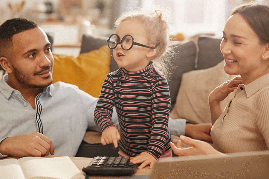 Portrait of happy young family counting home finances with cute little girl wearing glasses in sunlit apartment