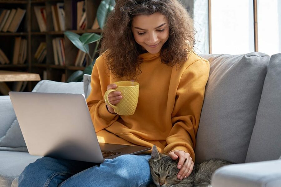 Happy relaxed hispanic teen girl student or freelancer relaxing sitting on couch holding laptop on lap working studying from home drinking tea, playing with cute pet cat in cozy sunny room on sofa.