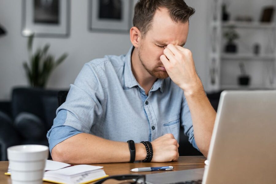 Overworked young entrepreneur in tired from work with a laptop, holds on to the bridge of the nose his eyes closed. Stressed guy works remotely at home feels eyes strain
