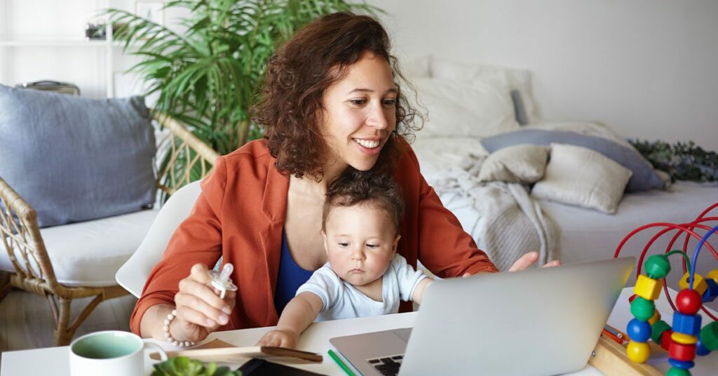 woman working at desk at home using laptop, holding baby on her lap