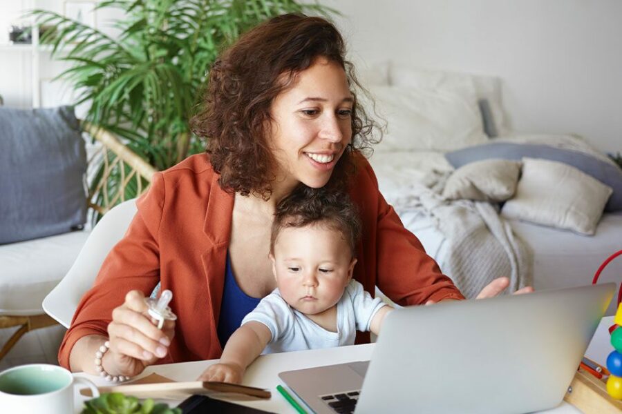 woman working at desk at home using laptop, holding baby on her lap