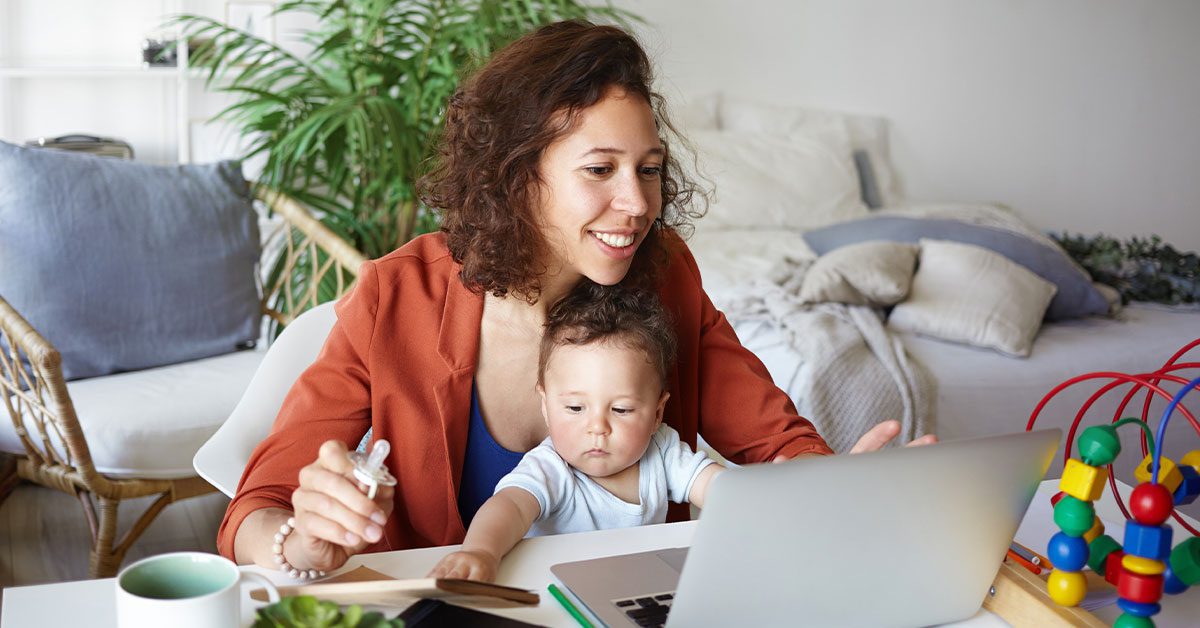 woman working at desk at home using laptop, holding baby on her lap